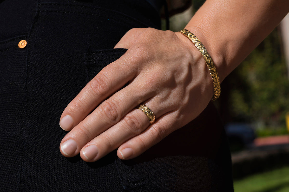 Hand wearing a 6mm gold Hawaiian engraved band and bangle against a dark background