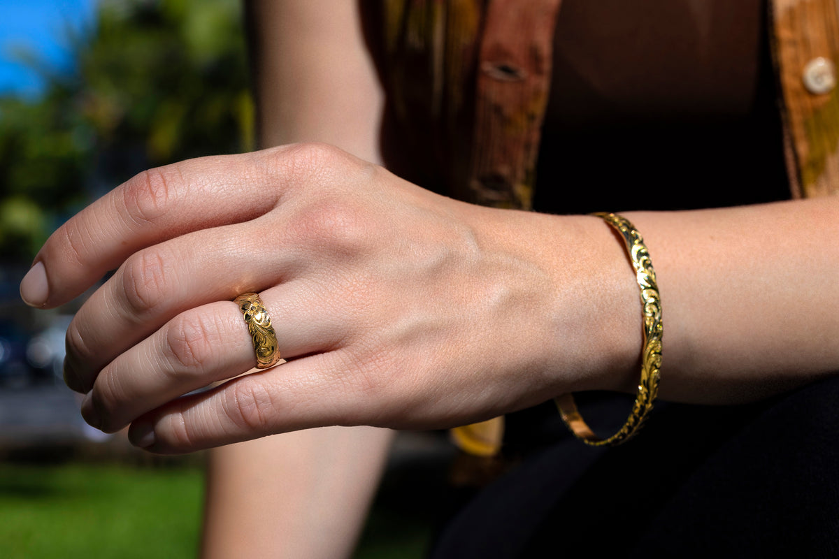 Woman's hand wearing a 6mm yellow gold Hawaiian engraved wedding band with a matching gold bangle