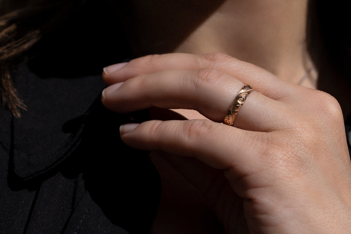 Close up of hands with a slim gold Hawaiian engraved ring against a dark background