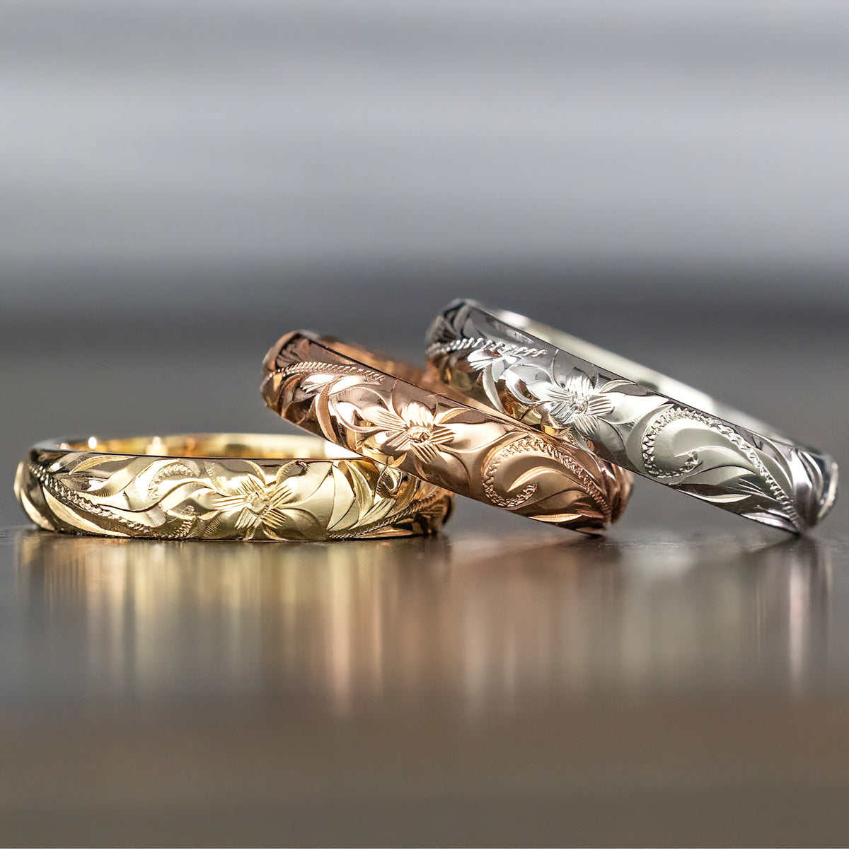 Three 4mm Hawaiian heritage rings in rose, yellow and white gold on a dark surface