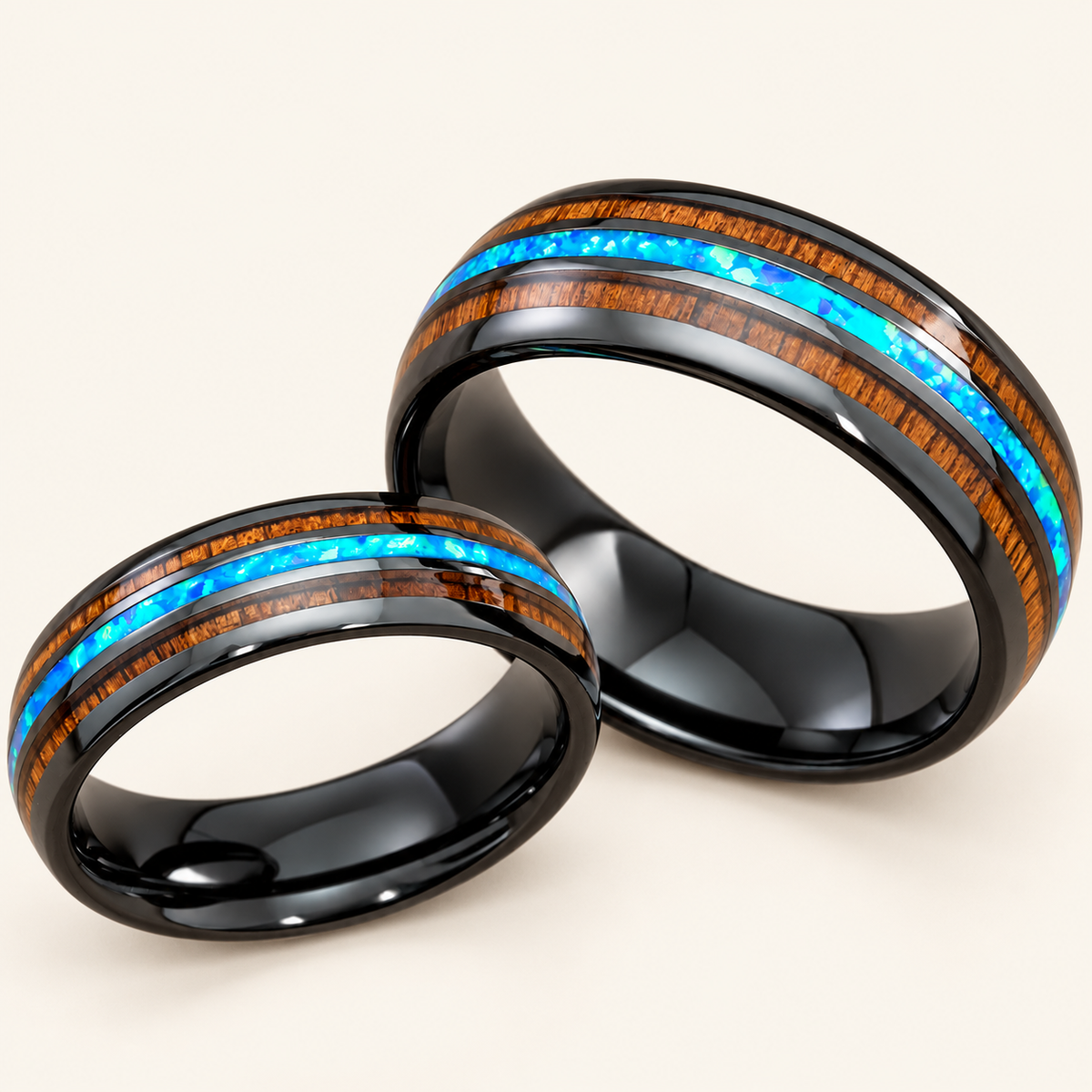 Pair of 6mm and 8mm black ceramic rings with blue opal center inlays and Hawaiian koa wood stripes, on an ivory background