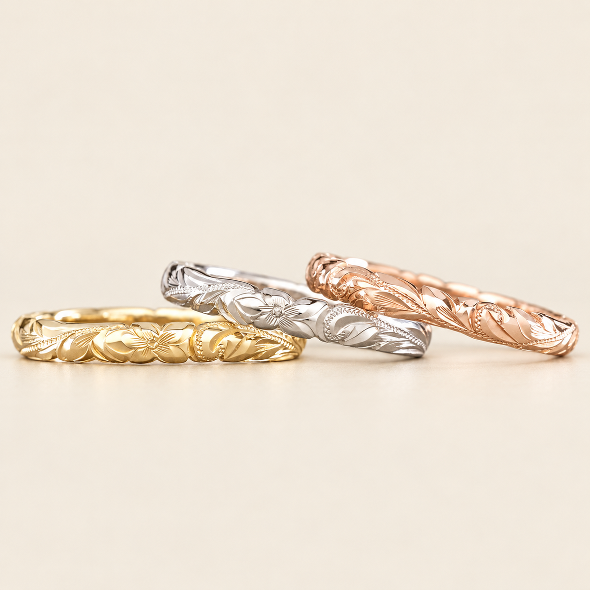 Three slim 3mm Hawaiian wave-edge rings in yellow, white and rose gold with hand-engraved plumeria, leaves and scrolls, shown on ivory.
