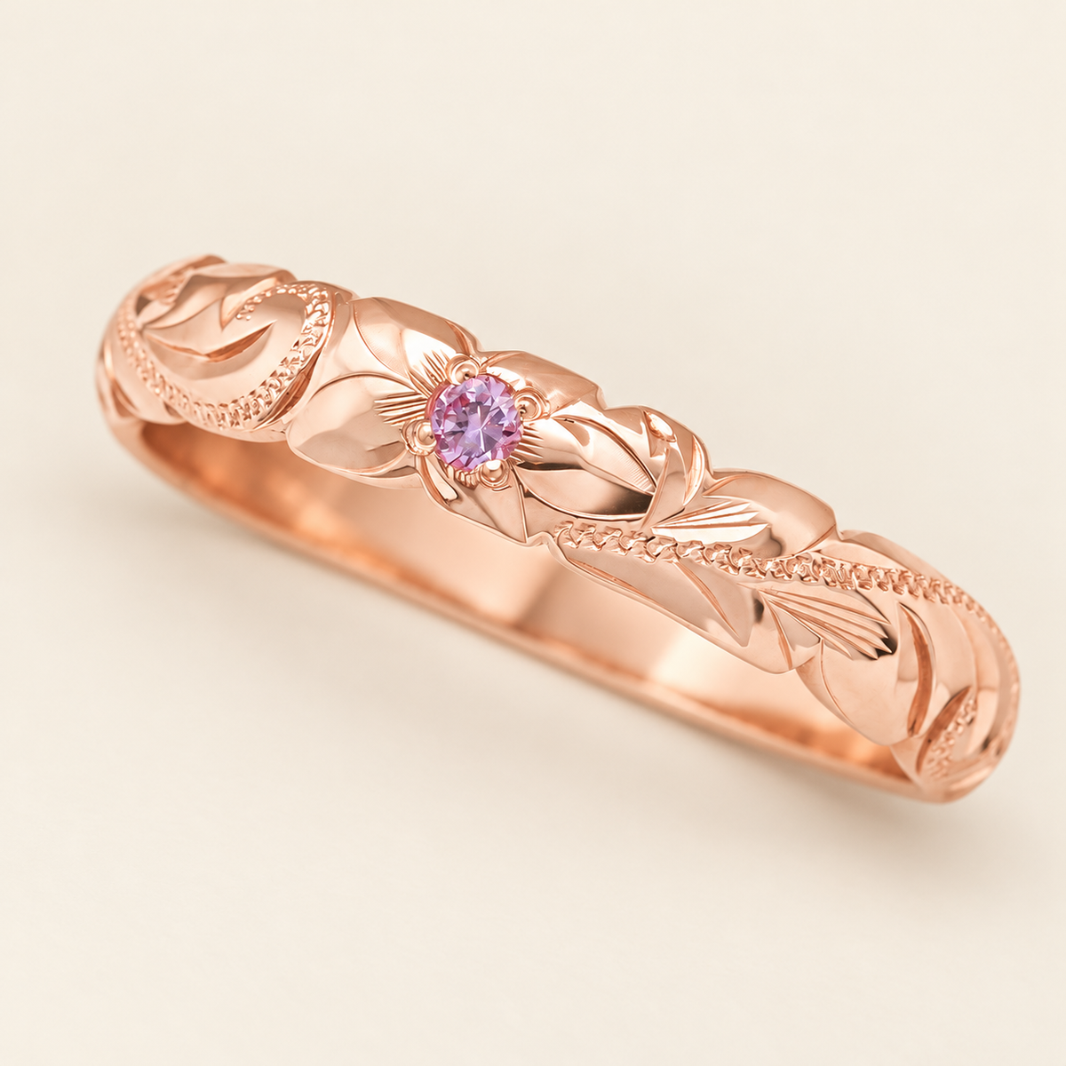 Rose gold Hawaiian ring with a pink sapphire in a four-prong floral setting, leaf and scroll engraving, on ivory.
