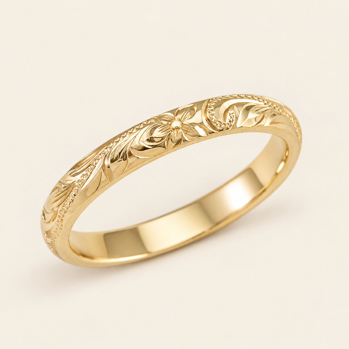 Slim yellow gold Hawaiian ring with a hand-engraved flower, scrolls and leaves, shown diagonally on ivory.