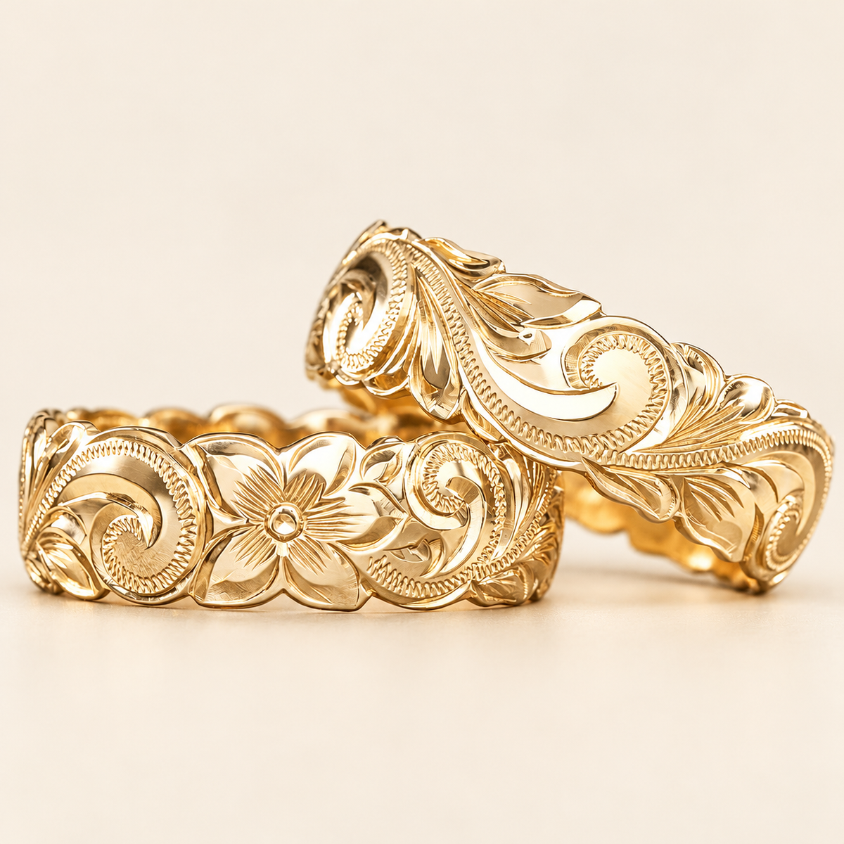 Two yellow gold 8mm Hawaiian rings with domed profiles, wave edges and engraved flowers, leaves and scrolls, shown on ivory.