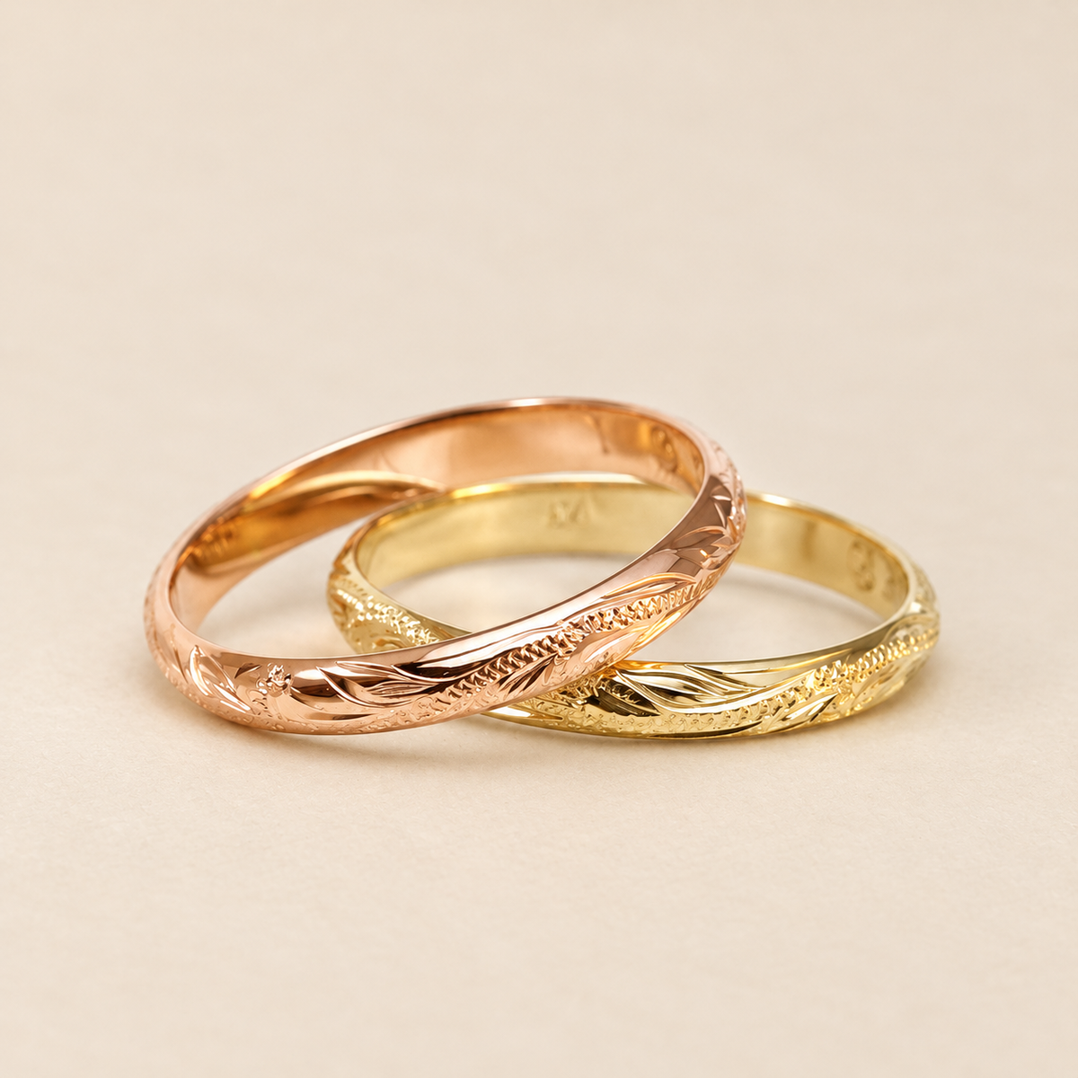 Rose and yellow gold 2mm Hawaiian hand-engraved rings overlapping on an ivory background.