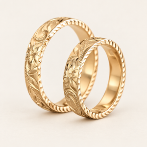 Two solid gold 4mm Hawaiian hand-engraved rings standing upright, with flower and scroll designs and diamond-cut edges on an ivory background